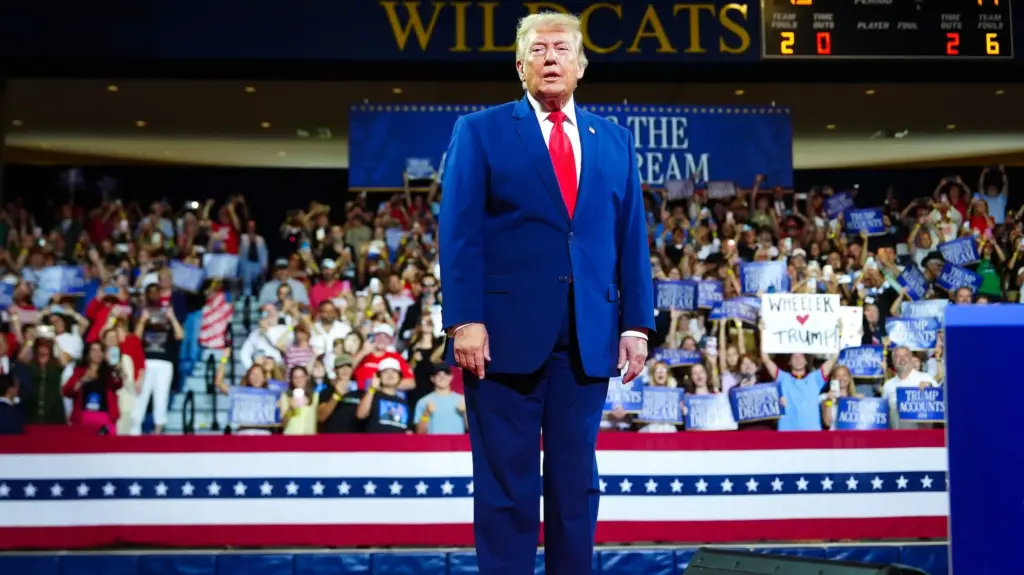 Donald Trump in a blue suit and red tie at a rally in an indoor stadium.