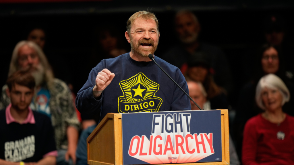Graham Platner speaks into a microphone behind a podium with a sign reading