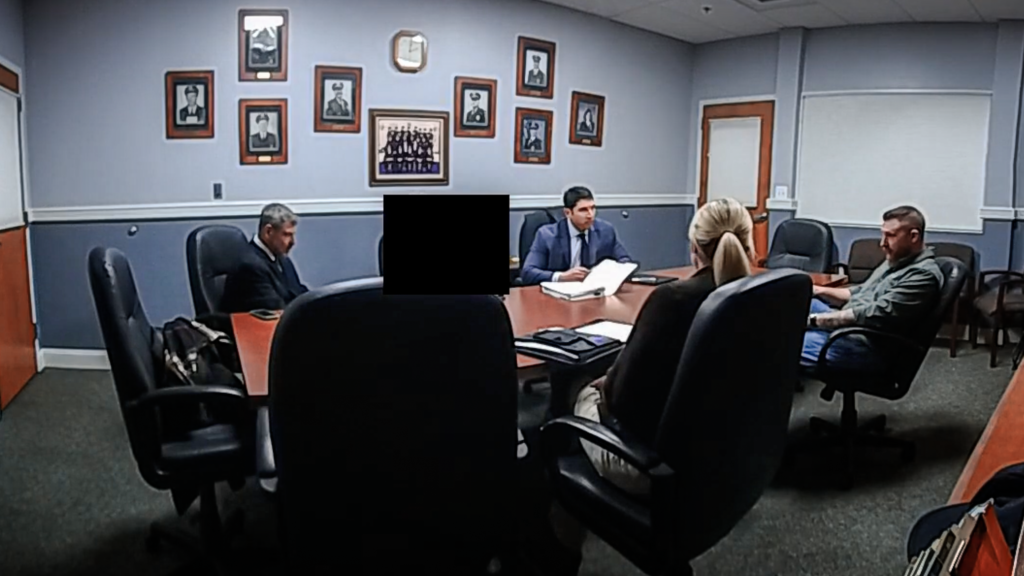 A video still of several people sitting in spacious black chairs around a brown table in a conference room with blue walls. On one wall hangs half a dozen photos of police officers. The identity of one person sitting at the table is obscured with a black rectangular box.