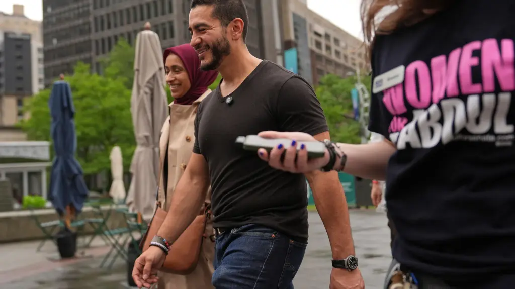 Abdul El-Sayed walks with his wife, Sarah Jukaku, past some picnic chairs and umbrellas in an outdoor city area. Another person holding a phone in their right hand and wearing a