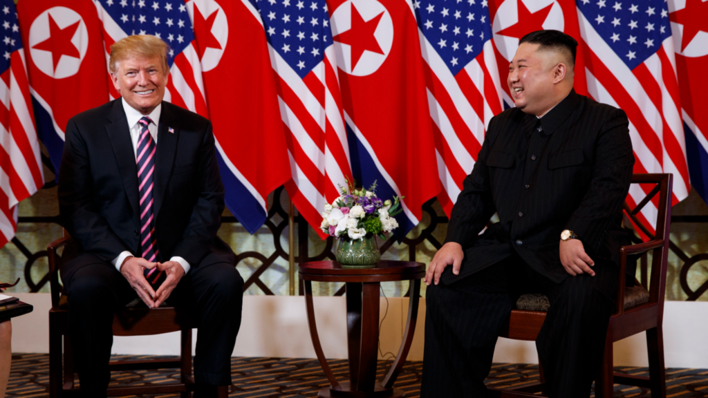 Donald Trump is smiling straight at the camera (left) while Kim Jong Un (right) smiles while looking at Trump. Both are sitting in wooden chairs indoors and several US and North Korea flags are in the background.