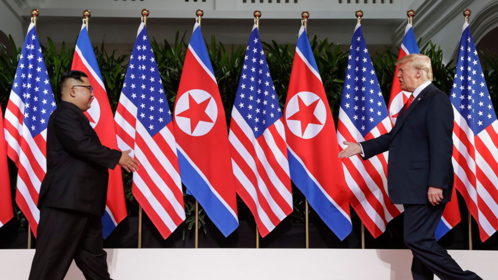 Donald Trump (right) and Kim Jong Un (left) walk toward each other. Both have their right arms outstretched to shake each others' hand. Alternating American flags (5) and North Korean flags (4) are in the background.