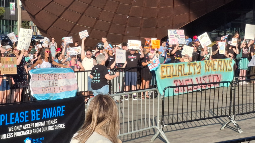 An individual wearing a black shirt and blue pants walks down a line of rallygoers hold signs supporting trans rights and inclusion in sports.
