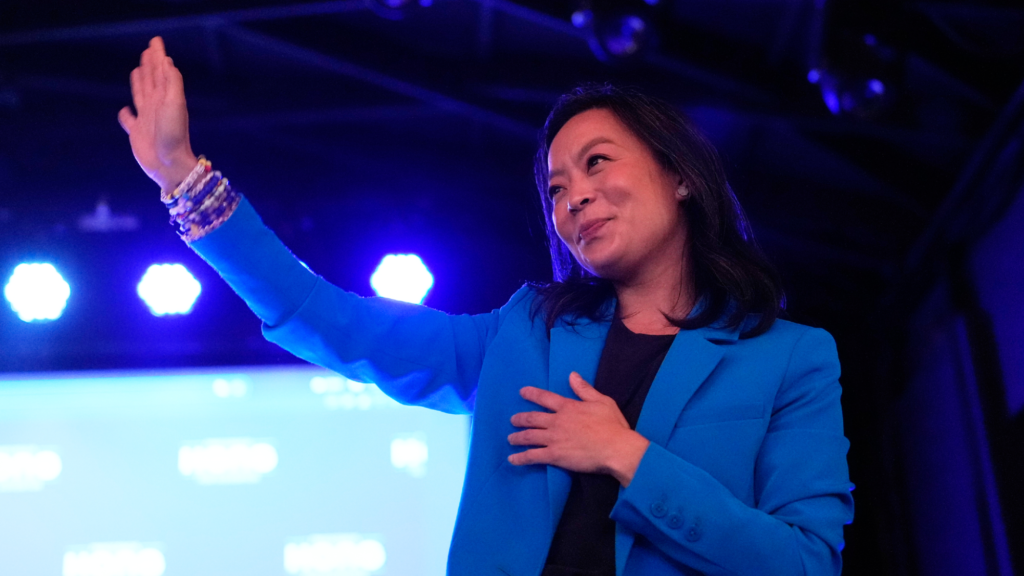 Wisconsin Democratic gubernatorial candidate Francesca Hong waves to supporters (who are not shown) with her right hand. Her left hand is over her heart. Hong is wearing a blue blazer and several bracelets on her right wrist. She is smiling.