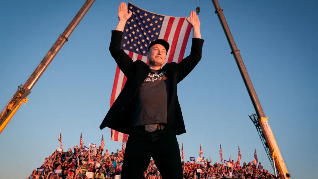 Elon Musk leaps into the air with arms raised above his head, with a slight smile on his face. Musk is on a stage in front of bleachers full of people. Outdoors under a blue sky and setting sun, two cranes hoist a huge American flag overhead.