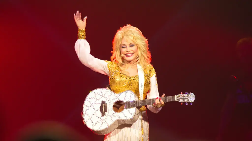 Dolly Parton waves to the crowd from a stage bathed in red light, smiling in a gold sequined top with sheer white sleeves and holding a rhinestone-studded white acoustic guitar.