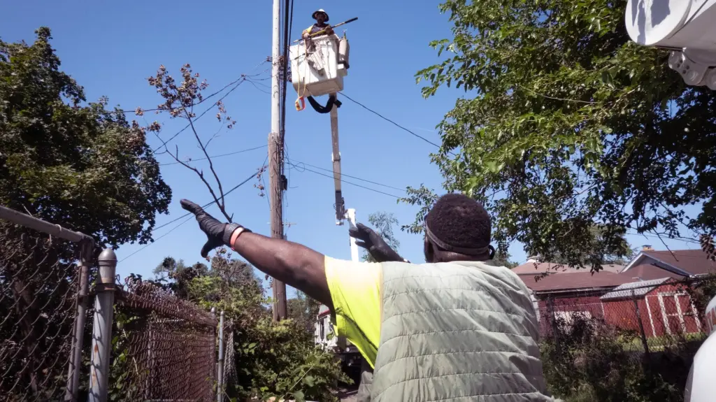A black man seen from behind is waving at a utility worker in a cherry picker on a sunny day