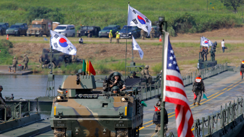 A military tank painted with camouflage colors is stationed on a bridge. Three soldiers are visible in the vehicle. In the background are more soldiers and several South Korean flags. In the foreground is the American flag. This is a joint military training drill with both South Korean and US forces.