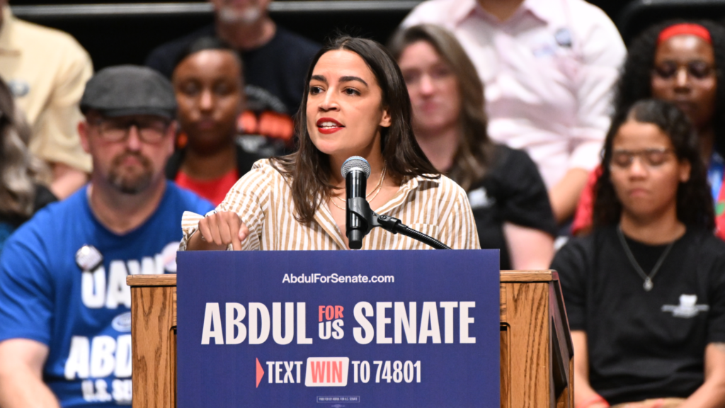 U.S. Rep. Alexandria Ocasio-Cortez stands at a brown wooden podium and speaks into a microphone. A sign in front of the podium reads in part