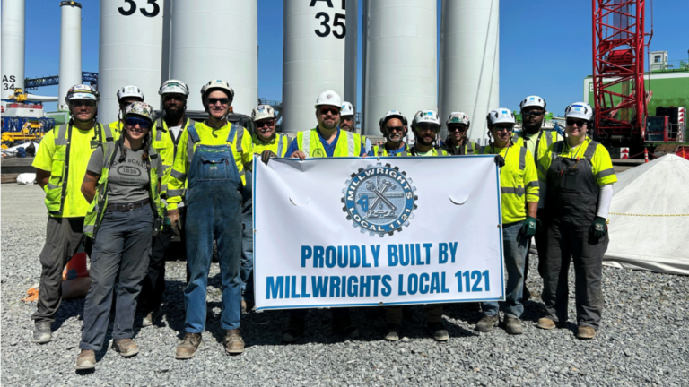 A bunch of workers in bright yellow shirts and white hardhats, many in overalls, pose with a union banner in front of a bunch of turbine shafts.
