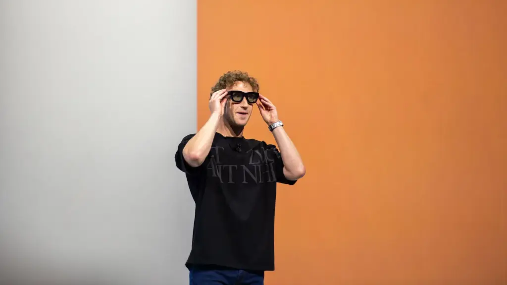 Mark Zuckerberg, a white man in a large black t-shirt, in front of an orange and white backdrop, wearing large black Meta glasses.