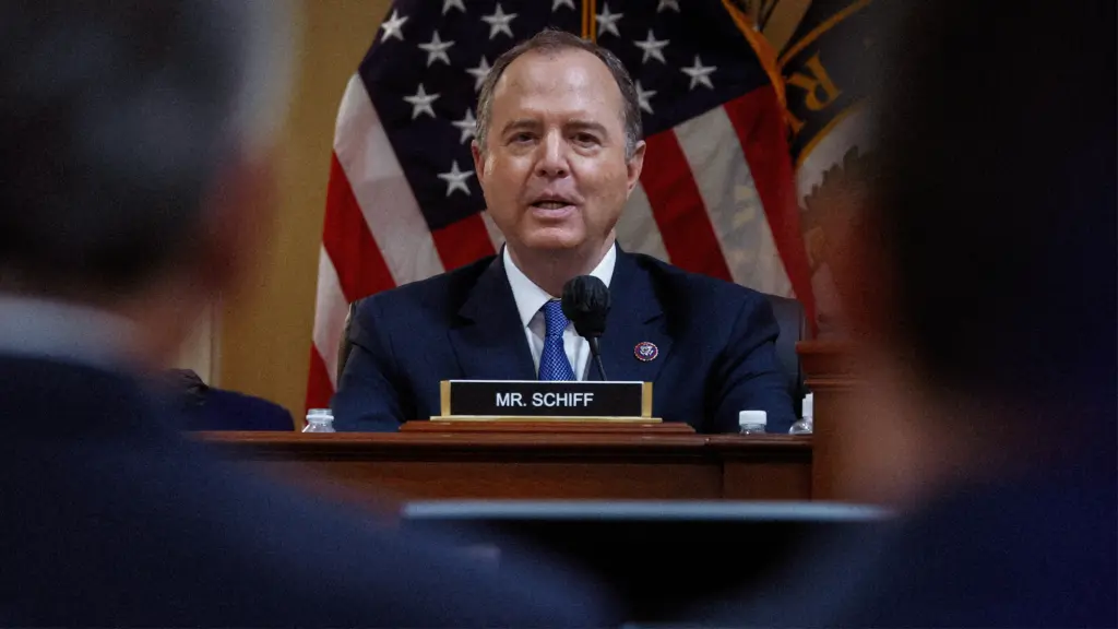 A man in a suit jacket and tie speaks into a microphone. He is seated behind a dais that has a nameplate that reads “Mr. Schiff.” Behind him is the American flag. In front of him, to the left and right, are two men, also in suits, watching Schiff as he speaks.