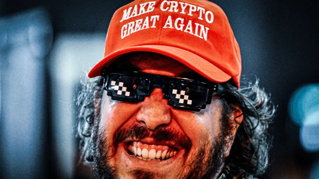 A closely cropped photo of the face of a middle-aged man with a beard, smiling broadly as he wears a red baseball cap that reads “Make Crypto Great Again.” He also wears sunglasses associated with the “Deal With It” meme.