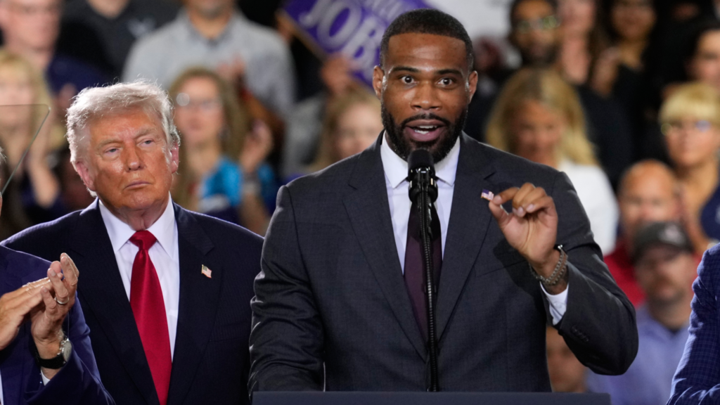 Amir Hassan, dressed in a suit, speaks into a microphone as he gives a speech at a campaign event. Standing to his right and slightly behind is President Donald Trump. We also see over a dozen people behind the pair in the crowd listening