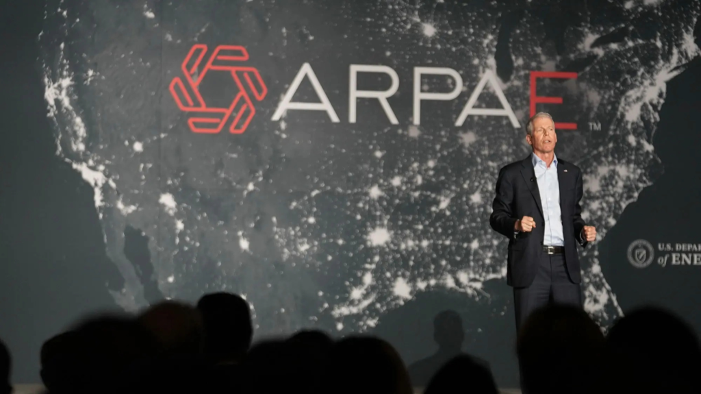 A man in a black suit speaks on stage with a U.S. map, ARPA-E text, and the Department of Energy logo on the backdrop behind him.