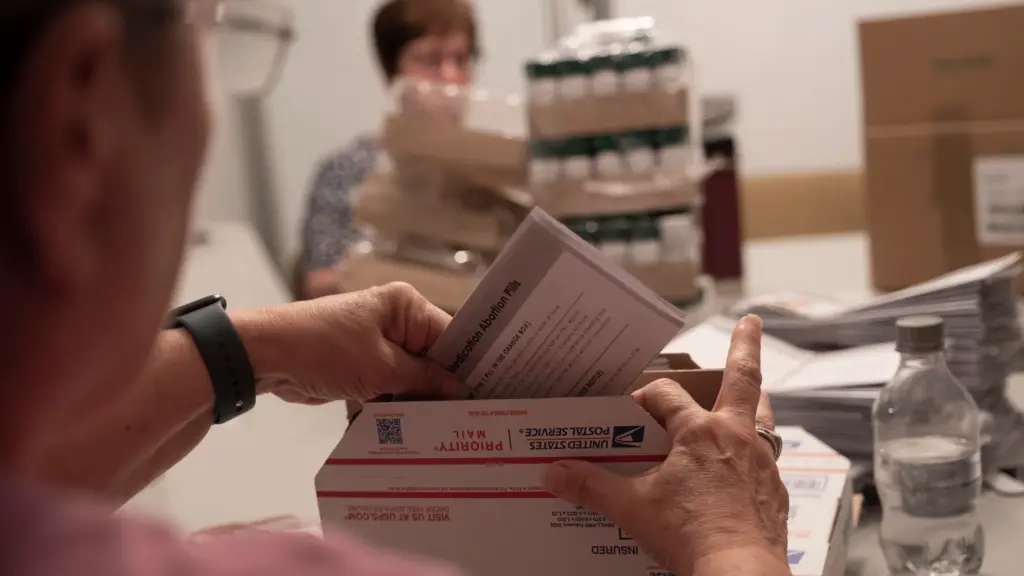 A person seen from behind slips a folded sheet of paper that reads in part “Medication Abortion Pills” into a United States Postal Service box. Out of focus and partially obscured, a woman with short hair sits on the other side of a table loaded with stacks of paper and shrink-wrapped cases of pills.