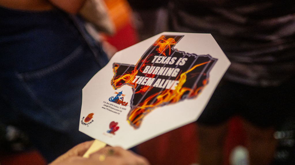A hand holding a sign with prison cell bars and fire in the shape of the state of Texas. It reads