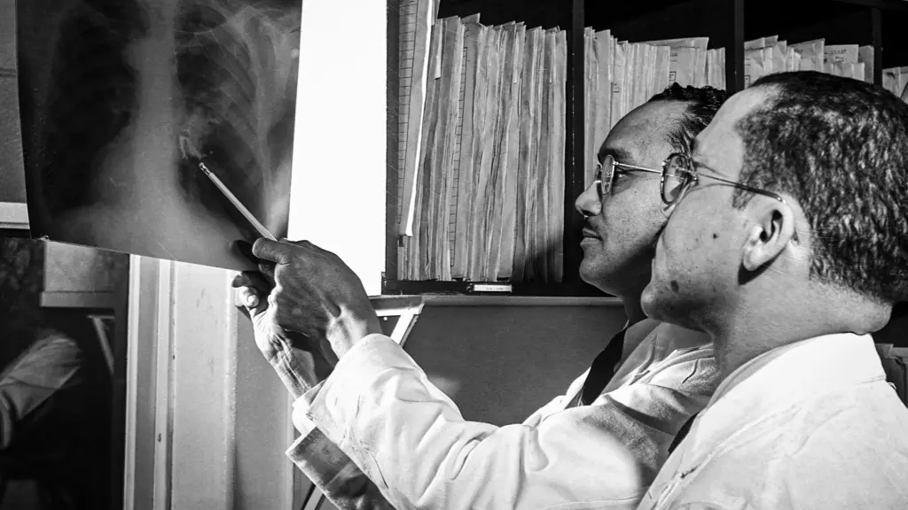 In a black-and-white photo, a Black male doctor in a white coat and glasses uses a pencil to point at an X-ray, which he holds up to light with the other hand. Beside him is a Black male intern, also dressed in a white coat and glasses.