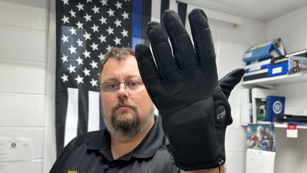 A man holds up a shock glove in front of a blue lives matter flag.