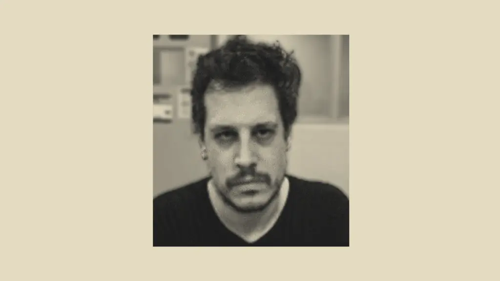 A booking photograph of a middle-aged man with untrimmed facial hair and a disheveled head of dark hair staring fiercely as he is being processed by law enforcement.
