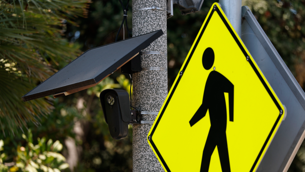 A photo shows A Flock camera mounted to a light pole. The camera is black and has wires traveling up to a small black solar panel. There is a yellow pedestrian walking sign in front.