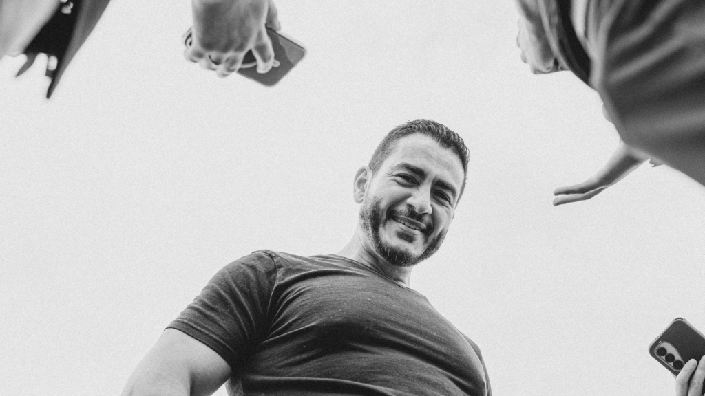 A black-and-white photo of a middle-aged man with dark hair and a dark beard smiling as he looks down toward the camera. Framed against a clear sky, he dressed in a black t-shirt. Around him, the hands and smartphones of reporters used as recording devices can be seen in the frame.