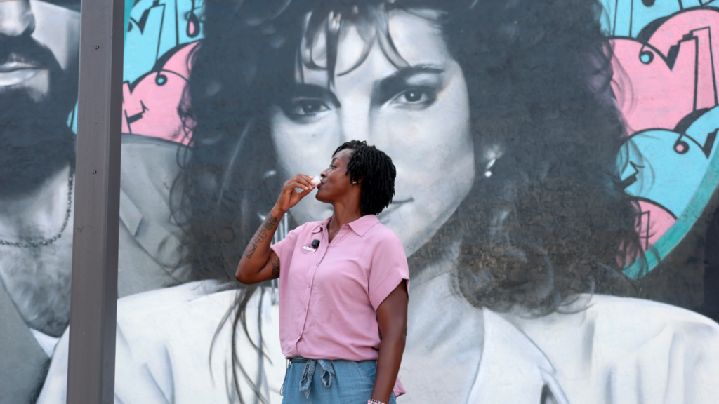 Angie Nixon is wearing a pink shirt and is holding a small cup to her mouth with her right hand. Behind her is a mural of singer Gloria Estefan in black and white.