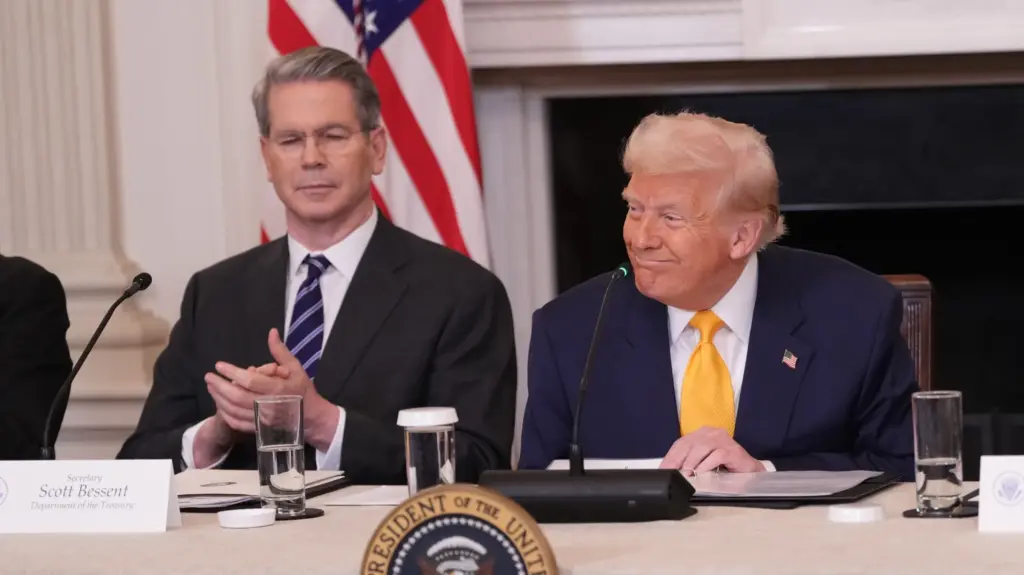 Donald Trump, seated, smiles behind a presidential seal and a tabletop microphone. He is wearing a navy suit jacket and gold-colored tie. To his right sits Treasury Secretary Scott Bessent, who is clapping and wearing a dark suit jacket and blue striped tie. Behind them a US flag stands.