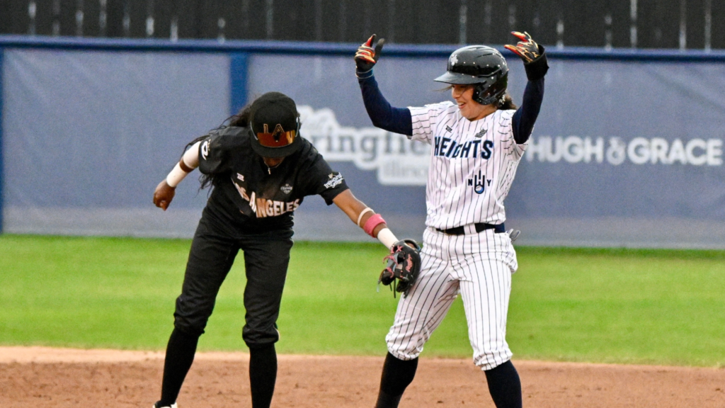 A Los Angeles Queens baseball player dressed in a black jersey tags a New York Heights player in a white jersey with blue stripes. The Heights player is celebrating because she is already on-base and safe.