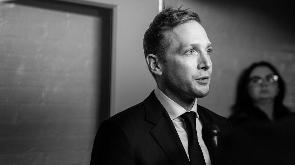 A black-and-white portrait of a man in a suit and tie speaking into a microphone, with a woman wearing glasses visible in the blurred background.