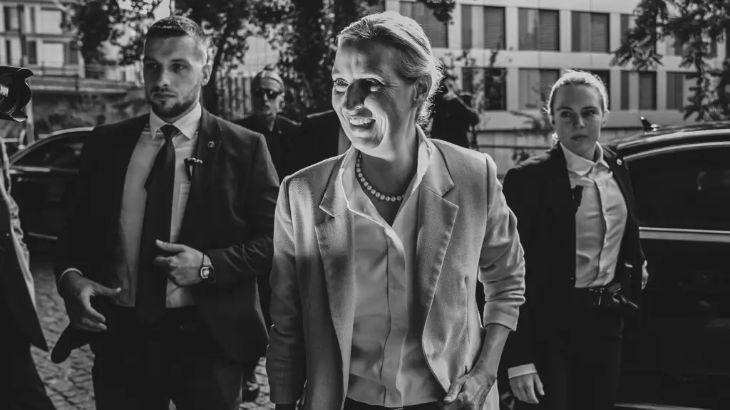 A black-and-white photograph of a woman dressed in light colors smiling as she is is lit brightly by a flash camera. Guards, a man and a woman, dressed in black suits and white shirts stand behind her.