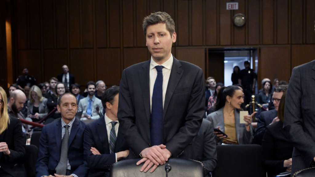 Sam Altman is wearing a black suit and is standing behind a black chair. His hands are folded and are resting on top of the chair and he is looking slightly down. Dozens of people sit behind him in a room during a Senate committee hearing.