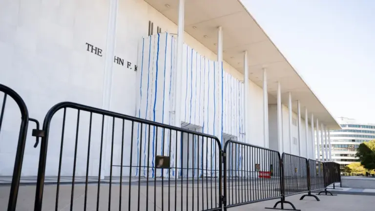 A fence stands in front of the facade of the Kennedy Center, which is partially covered by a white and blue tarp obscuring the center's name.