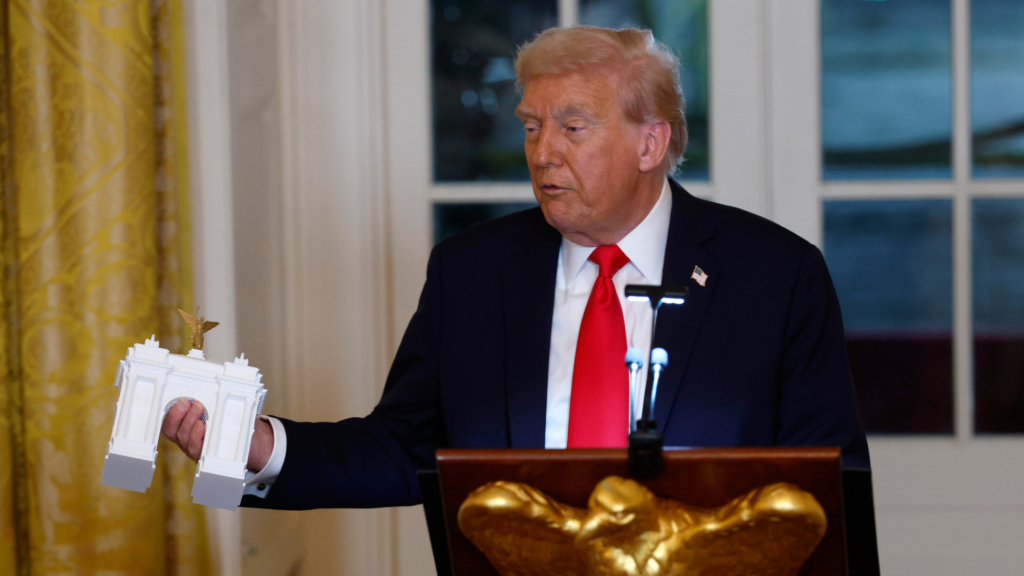 Donald Trump is standing at a podium and speaking into a microphone. He is looking to his right and is holding a model of a white arch that he wants to build in D.C. in his right hand.