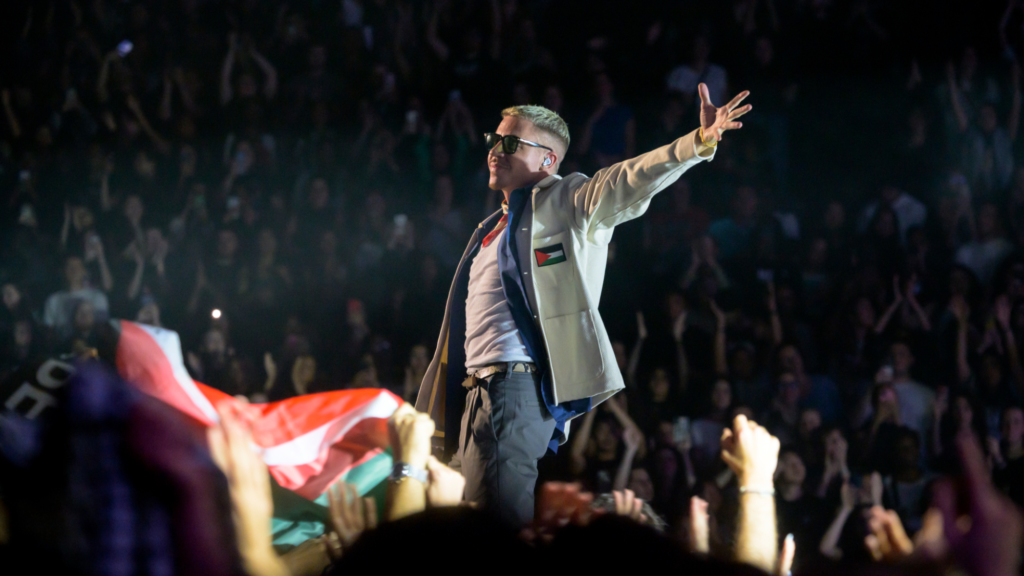 Macklemore is standing onstage during a concert. He is wearing a white jacket with a Palestinian flag on the left chest side. A crowd of audience members are shown in both the foreground and background.