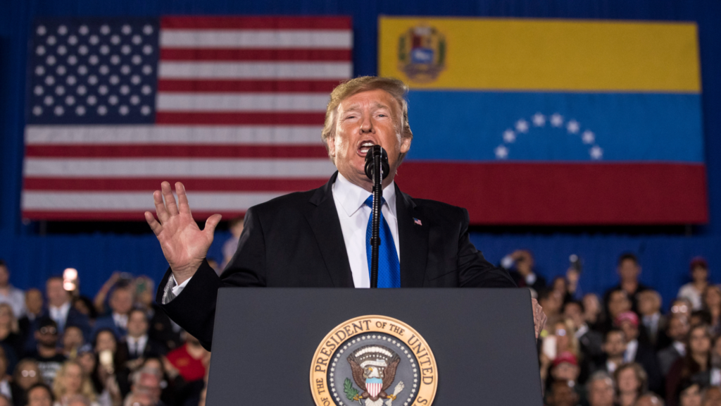 Donald Trump is wearing a black suit and blue tie. He is speaking into a microphone behind a podium. Behind him, dozens of audience members are seen listening. Above the audience is the US and Venezuela flags.