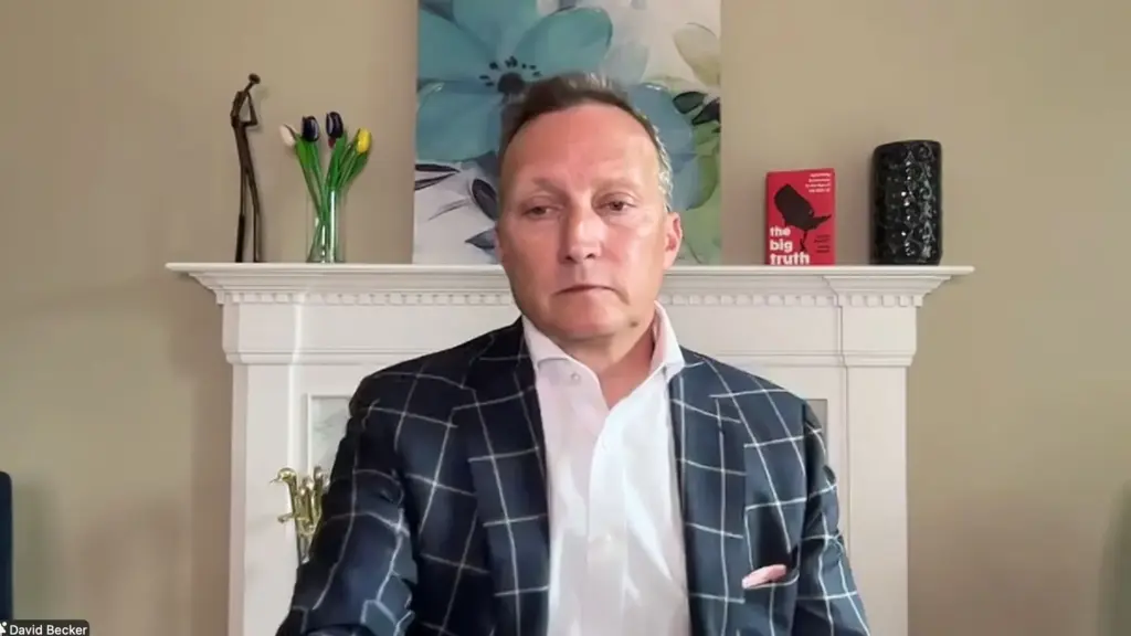 A white man in a blazer and white shirt sits in front of a white painted fireplace with a painting and some things on the mantle