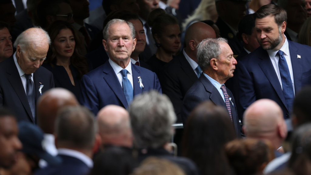 Four men are in focus in the photo (Left to right): Joe Biden, George Bush, Michael Bloomberg, and JD Vance. Several people are seen closer to the camera facing them. Everyone is standing up.
