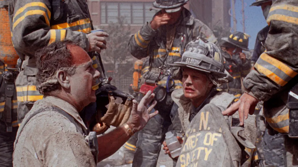 Seven firefighters, some sitting and some standing, talk emotionally as they stand amid rubble. Their uniforms are caked with gray dust, and one man in the foreground has the dust on his face.