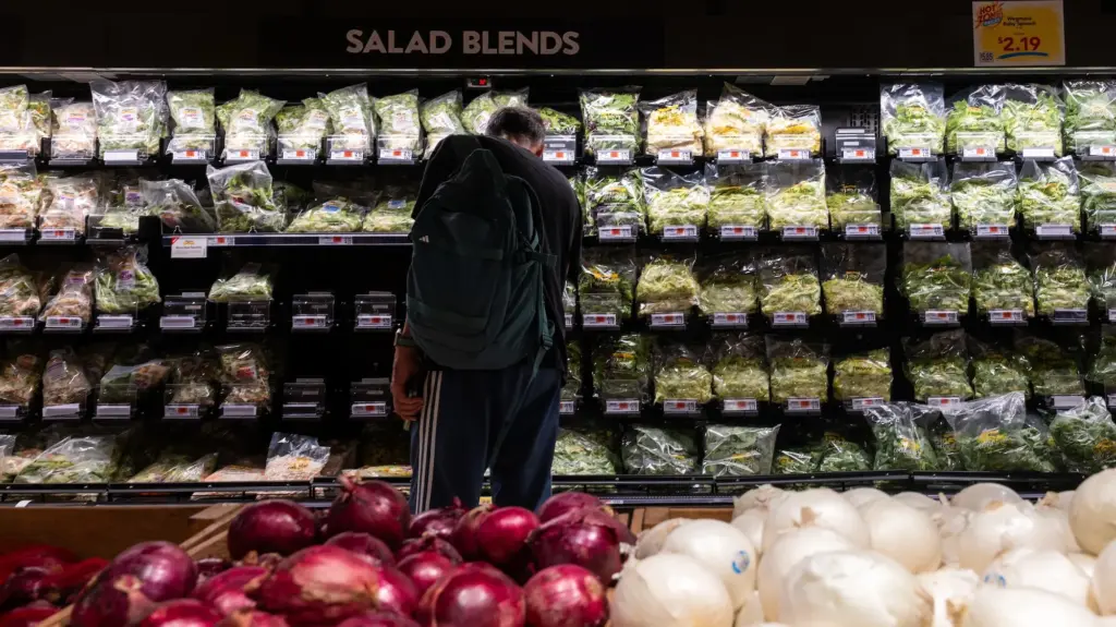 A man wearing a backpack looks at varieties of salad blends available in a grocery store's produce section.