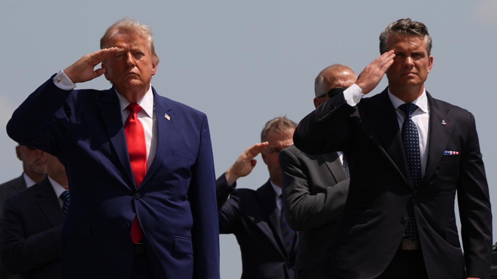 Donald Trump and Pete Hegseth, both dressed in dark suits are saluting with their right hands while standing outside. Four people are seen standing behind them.