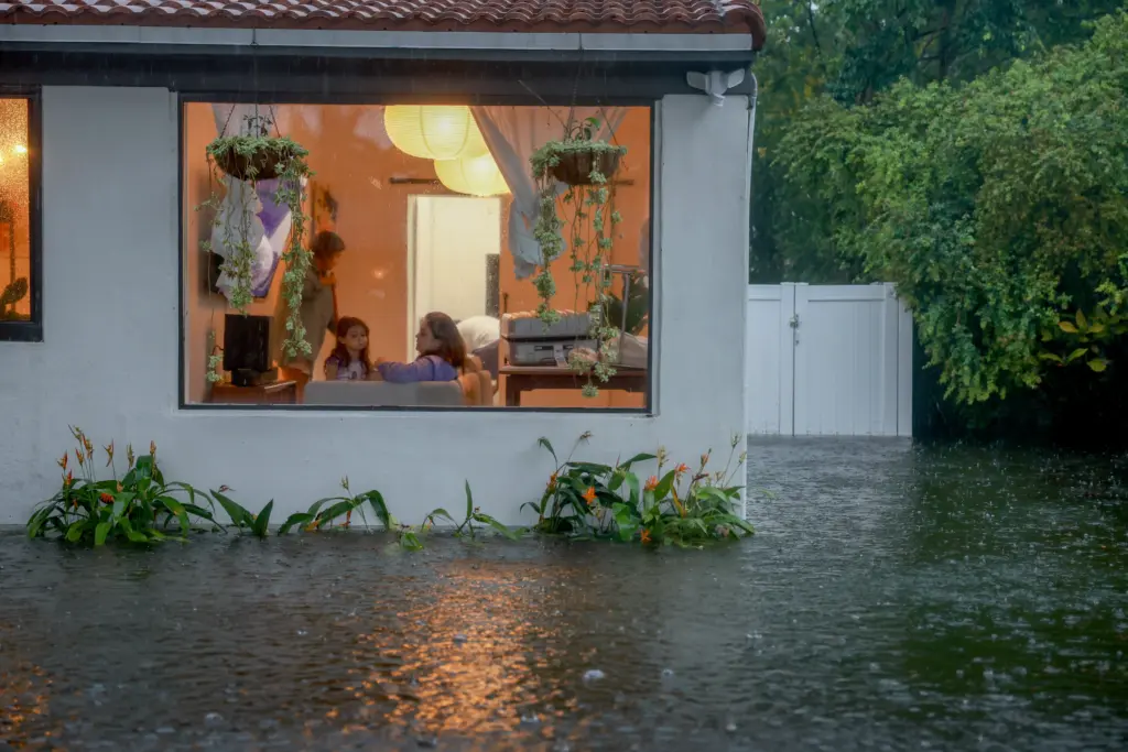 In the upper left, a window into a well-lit room with plants and a seemingly-packed bag. There are two adult females, and a young girl looks out the window. All around, there is water flooding the area and persistent raindrops.