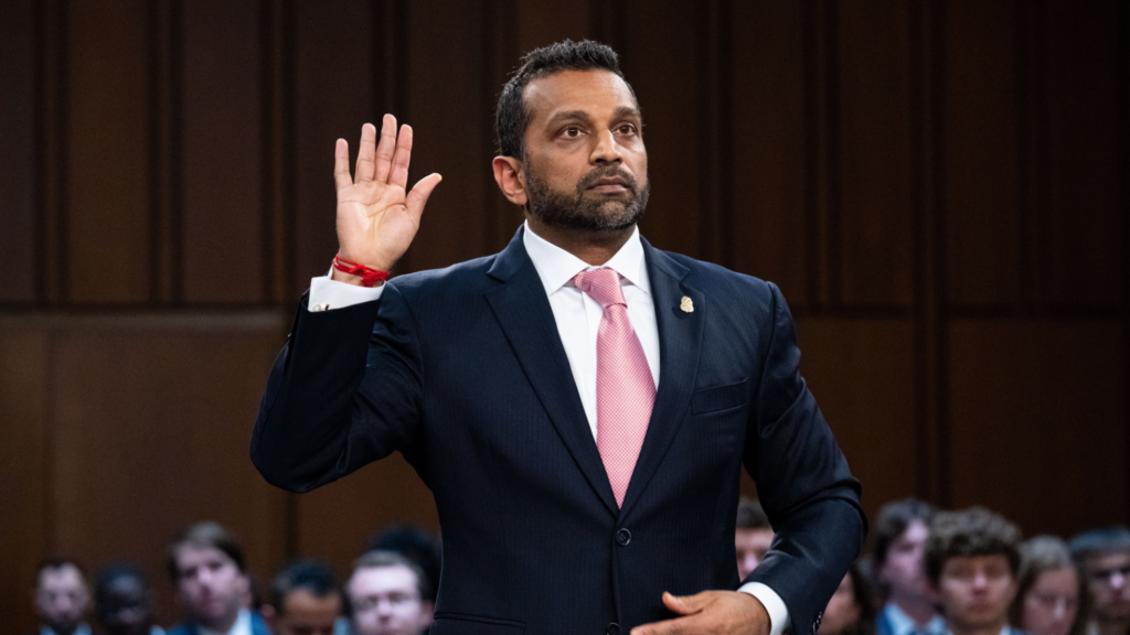 Kash Patel is sworn in to a Senate hearing. He is wearing a dark suit and is raising his right hand with his palm facing forward. His left hand is holding his stomach. Over a dozen people are seen sitting behind him dressed in formal wear.
