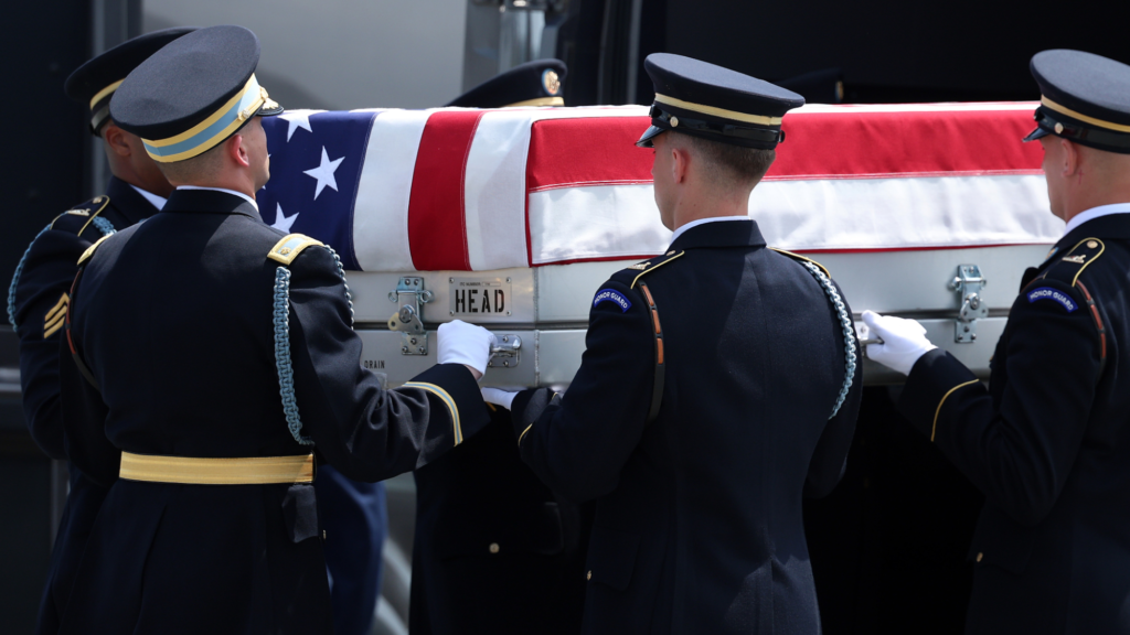 Five people dressed in US military attire are seen holding a transfer case draped in the US flag. The case itself is silver.