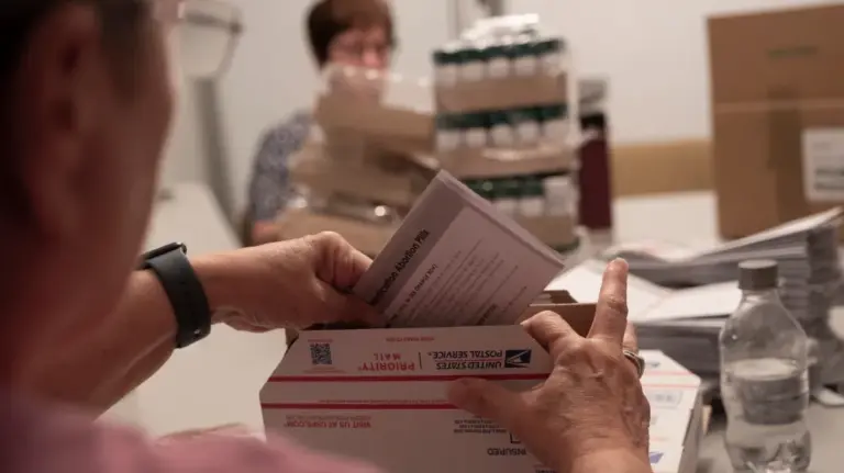 A person seen from behind slips a folded sheet of paper that reads in part “Medication Abortion Pills” into a United States Postal Service box. Out of focus and partially obscured, a woman with short hair sits on the other side of a table loaded with stacks of paper and shrink-wrapped cases of pills.