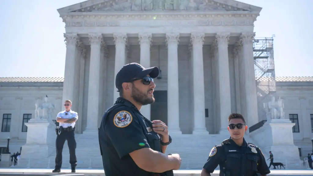 A white man with a beard and sunglasses and a blue uniform stands outside the supreme court building on a sunny day with two others.