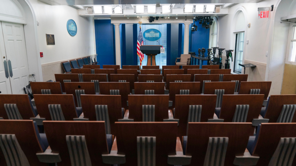A photo at the back of the White House Press Briefing Room. The room does not have any people—just chairs, the podium, and other furniture. The headlights on the ceiling are turned on.