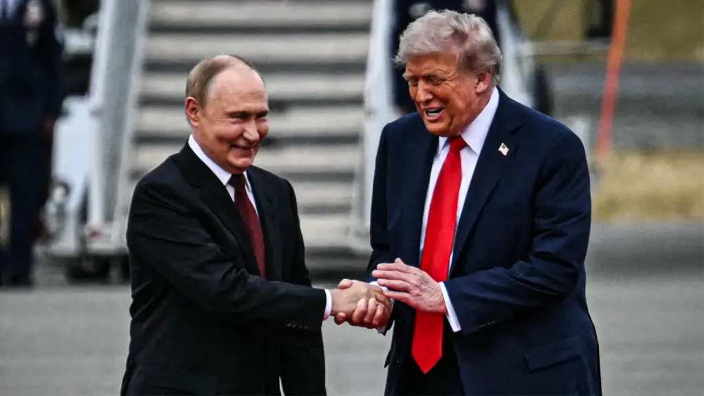 A photograph of two older white men, both wearing a dark suit and red tie, shaking hands warmly with expressive smiles. Donald Trump, on the right, raises his left hand as if he’s about to pat Vladimir Putin’s right hand as he grips it.