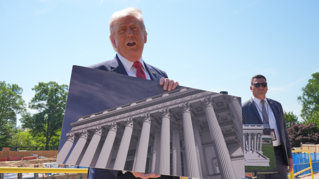Donald Trump holds a poster the size of his upper body of a rendering of the outside of the White House ballroom. He is outside at a construction site. Another person in a suit with sunglasses is standing behind Trump.