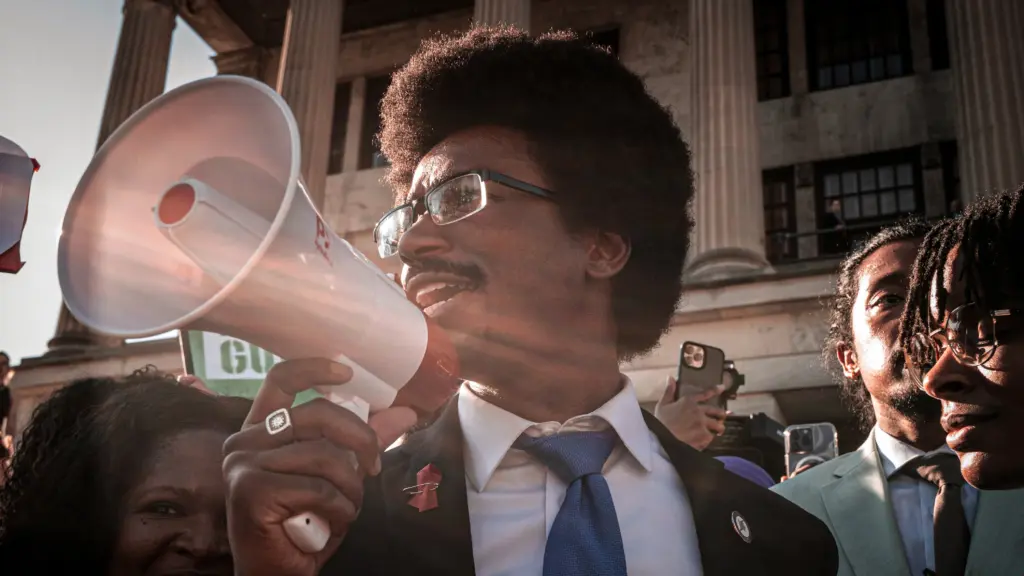 A young Black man with a trimmed Afro hairstyle and a mustache, wearing glasses and a suit with a white shirt and blue tie, speaks into a bullhorn. He is surrounded by other people, mostly Black, as they stand on the steps of a neoclassical building with tall columns.
